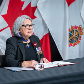 La gouverneure générale Mary Simon est assise à une table. Devant elle il y a un cahier ouvert et derrière elle on peut voir le drapeau canadien et le drapeau des Forces armées canadiennes.