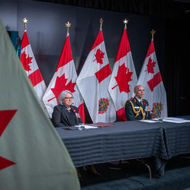 Governor General Mary Simon is sitting at a long table with General Wayne Eyre. They are both facing the camera. Behind them are Canadian and Canadian Armed Forces flags. 