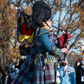 A bagpiper plays the bagpipes by the National War Memorial. They are wearing a bearskin hat, and a plaid military uniform. Veterans can be seen in the background.
