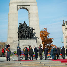 Une file composée de plusieurs personnes fait face au Monument commémoratif de guerre du Canada. La gouverneure générale Mary Simon et M. Whit Fraser se tiennent parmi eux. Le ciel est bleu.