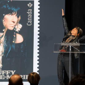 Buffy Sainte-Marie speaks at a podium. She is celebrating with her arms raised. A photo of her new commemorative stamp is visible to her right.