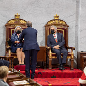 Leurs Excellences sont assises à une paire de trônes. Le secrétaire du gouverneur général lui remet un classeur contenant le discours du Trône.