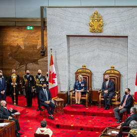 Vue large de la salle du Sénat. Leurs Excellences sont assises dans une paire de sièges trônes, le premier ministre est à la droite de Son Excellence. Le juge en chef du Canada est assis face à leurs Excellences.