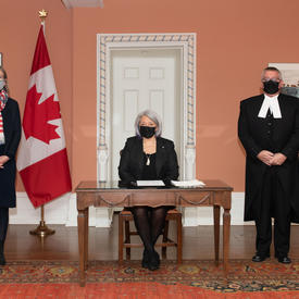 La gouverneure générale se tient entre quatre personnes. Toutes les personnes portent des masques. Un drapeax du Canada se trouve à l’arrière-plan.
