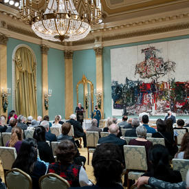 Vue de la salle de bal de Rideau Hall. La gouverneure générale Mary Simon se tient sur un podium à l'avant de la salle.