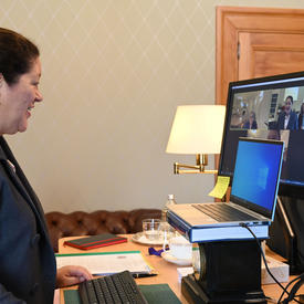 A woman participates in a virtual call on her computer. She is sitting at a desk with two monitors before her.