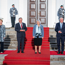 Their Excellencies and two other people are standing on the red carpet at the base of the stairs outside a large white building.