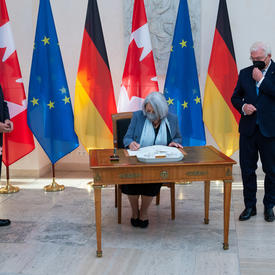 The Governor General is sitting at a wooden desk. She is signing the guest book. President Steinmeier is standing to her left. Ms. Elke Büdenbender and Mr. Whit Grant Fraser are standing to her right. There are several flags behind her. 