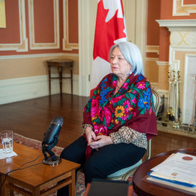 Governor General Mary Simon is sitting down. She is wearing a beautiful multi-coloured scarf. A Canada flag is behind her. A microphone is on a low table in front of her.