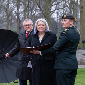 Her Excellency is walking in Green Park. A member of the military is holding a book in front of her. High Commissioner Ralph Goodale is to her right holding an umbrella.