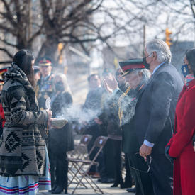 Governor General Simon is standing in a row of people. She is shrouded in smoke from a smudging ceremony. An Indigenous woman performing the smudging ceremony is standing before her.