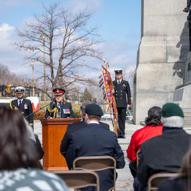 Governor General Simon is delivering remarks at the National War Memorial. People in various military uniforms are standing behind her. The photo is taken from the back of a crowd looking at her.