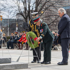 Governor General Simon is laying a wreath at the National War Memorial. Mr. Fraser and a man in a military uniform are standing beside her. There are rows of people sitting on chairs in the background.