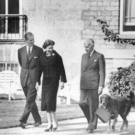 A black-and-white photo of The Queen and The Duke of Edinburgh walking outside Rideau Hall with Governor General Massey. A dog walks alongside them, carrying The Queen’s purse in its mouth. 