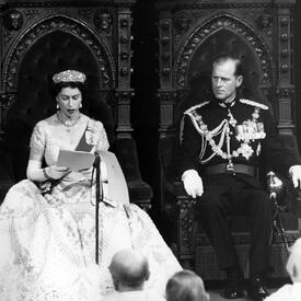 A black-and-white photo of The Queen and the Duke of Edinburgh seated in two thrones, in the Senate Chamber of Parliament. The Queen reads from a speech as the Duke looks on. They are both in formal attire.