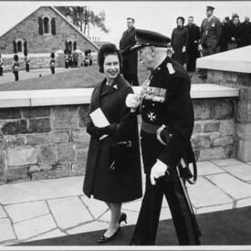 A black-and-white photo of The Queen walking with Governor General Vanier. They are walking along the stone ramparts of the Citadelle. A small crowd of people, guards in uniform and another stone building are in the background.