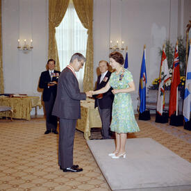 Jules Léger, dressed in a grey suit, shakes the hand of The Queen in the Ballroom at Rideau Hall. The Queen stands in front of him in a green dress and white heels. Three men look on in the background.