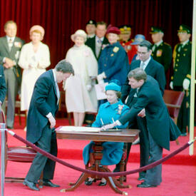 The Queen, dressed in a blue coat and hat, is seated at a wooden table. There is a large document on the table. Several politicians and others stand by watching. They are all standing on a large red carpet.