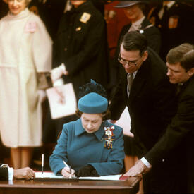 The Queen, dressed in a blue coat and hat, is seated at a wooden table. She is signing a document. Then-Prime Minister Pierre Elliott Trudeau is also seated at the table. Three other men look on.  