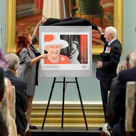 Le gouverneur général de l’époque, David Johnston, et Mme Siân Matthews, de Postes Canada, dévoilent un nouveau timbre en l’honneur de la Reine. Le timbre présente une image en couleur de Sa Majesté et une ancienne photo en noir et blanc de sa jeunesse.