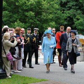 The Queen, in a matching blue coat and hat, walks along a pathway surrounded by trees on the Rideau Hall grounds. She is accompanied by the Secretary to the Governor General and by The Duke of Edinburgh. She smiles at a crowd of people. 
