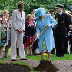 The Queen, in a matching blue coat and hat, shovels soil during a tree-planting ceremony, as a crowd of people look on. Stands of trees are in the background