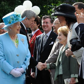 La Reine, qui porte un manteau bleu et un chapeau assorti, sourit à une foule. Derrière elle, le premier ministre de l’époque, Stephen Harper, serre la main d’une femme.