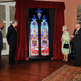 The Queen stands beside a stained glass window installment. A set of red curtains hangs on either side of the window. The Queen is accompanied by five other people. 