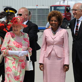 The Queen stands next to then-Governor General Michaëlle Jean and Jean-Daniel Lafond. They are outside an airport. An RCMP officer and other security personnel are behind them. 