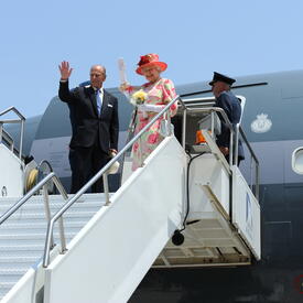 The Queen and The Duke of Edinburgh wave from atop a set of stairs connected to a plane. 