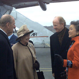 The Queen and The Duke of Edinburgh speak with Governor General Adrienne Clarkson and John Ralston Saul. They are standing on the tarmac of an airport, beside a plane. 