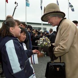 The Queen, dressed in a beige coat and matching hat, receives a bouquet of flowers from two children. A crowd of people stand behind them. Several flags on flagpoles are in the background.
