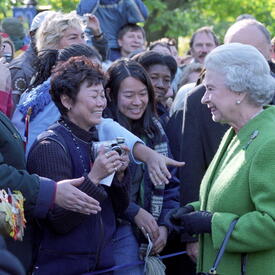 The Queen, in a bright green coat, smiles at a crowd of people standing outside. Several people are reaching forward to shake The Queen’s hand.
