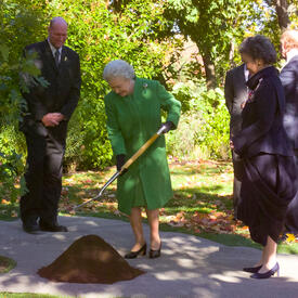 The Queen, wearing a green coat, holds a shovel above a small pile of dirt. Then-Governor General Adrienne Clarkson stands to her left. Greenery and trees are in the background.