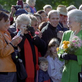  The Queen, holding bouquets of flowers and wearing a green coat, greets a crowd of people outside Rideau Hall.