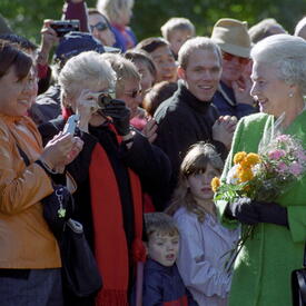 La Reine, qui tient des bouquets de fleurs et porte un manteau vert, salue une foule à l’extérieur de Rideau Hall.