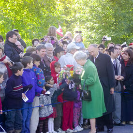 The Queen, holding bouquets of flowers and wearing a green coat, greets a crowd of people outside Rideau Hall.