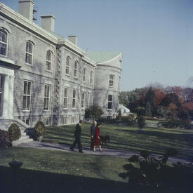 La Reine marche le long d’un chemin à l’extérieur de Rideau Hall avec le gouverneur général de l’époque, Vincent Massey. Des arbres aux couleurs d’automne sont à l’arrière-plan.