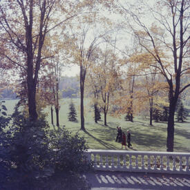 The Queen and then-Governor General Vincent Massey walk together across a lawn surrounded by trees in fall foliage.