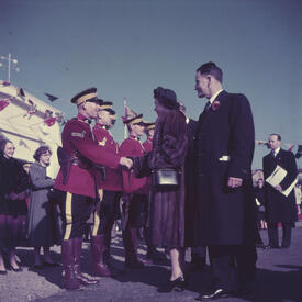 Princess Elizabeth, wearing a fur coat, shakes hands with an RCMP officer while three other officers stand at attention. The officers are wearing red dress uniforms. A ship and nautical flags are in the background.