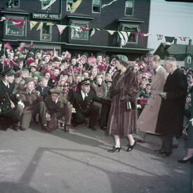 Then-Princess Elizabeth, wearing a long, fur coat, walks past a crowd of people. Several photographers are kneeling before her to take photos.