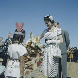 La Reine reçoit un bouquet de fleurs des mains d’une jeune fille autochtone en habits traditionnels. Le duc d’Édimbourg est derrière elle. On voit une foule à l’arrière-plan. 