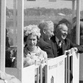 Une photo en noir et blanc de la Reine, portant un chapeau de fleurs, assise dans une nacelle en compagnie de Lester B. Pearson et d’un autre homme occupant les sièges derrière elle. On voit l’inscription «  Montréal 1967 » sur le côté de la nacelle.