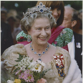 The Queen, holding a bouquet of flowers in her arms, smiles and poses. She is dressed in formal attire and wears the Order of Canada and other insignia. 