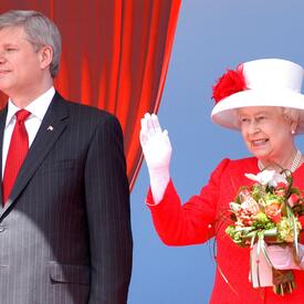  La Reine, vêtue d’une tenue rouge et d’un chapeau rouge et blanc, tient un bouquet d’une main et salue de l’autre. Elle sourit. Le premier ministre de l’époque, Stephen Harper, se tient à ses côtés, vêtu d’un complet noir et d’une cravate rouge. 
