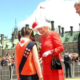  La reine Elizabeth sourit en recevant des fleurs d’une enfant qui porte un uniforme des Guides du Canada. Le premier ministre de l’époque, Stephen Harper, les regarde. On voit l’édifice du Parlement ainsi qu’une foule de personnes à l’arrière-plan.