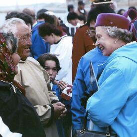 La Reine, qui porte un parka bleu et un chapeau rouge bordeaux, sourit à deux Aînés inuits en habits traditionnels. Un enfant tenant un petit drapeau du Canada regarde la scène. On voit une foule à l’arrière-plan.