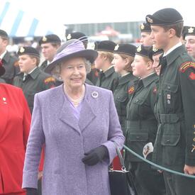 The Queen, wearing a purple coat and matching hat, walks past a group of cadets in uniform, standing at attention. 