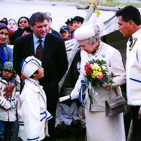 The Queen, dressed in white and carrying a bouquet of flowers, is greeted by a child wearing a white-and-blue ceremonial coat and hat. Two officials look on, while a crowd of people stand behind them. 