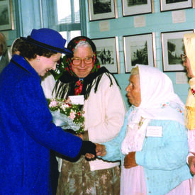 La Reine serre la main d’une femme portant un fichu sous le regard de deux autres femmes à la tenue similaire.